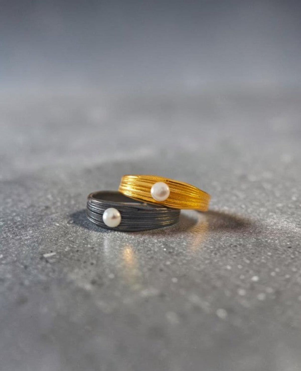 Two minimalist wire-wrapped rings on a textured gray surface. One ring is gold with a small white pearl, and the other is dark gray/black with a matching pearl, set against a dark gray gradient background.