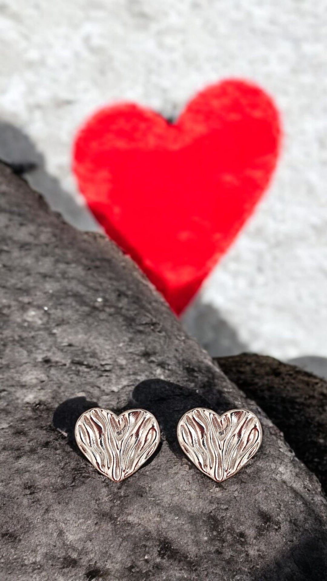 Sterling silver heart stud earrings with folded draped texture, photographed on rock with red heart graffiti background