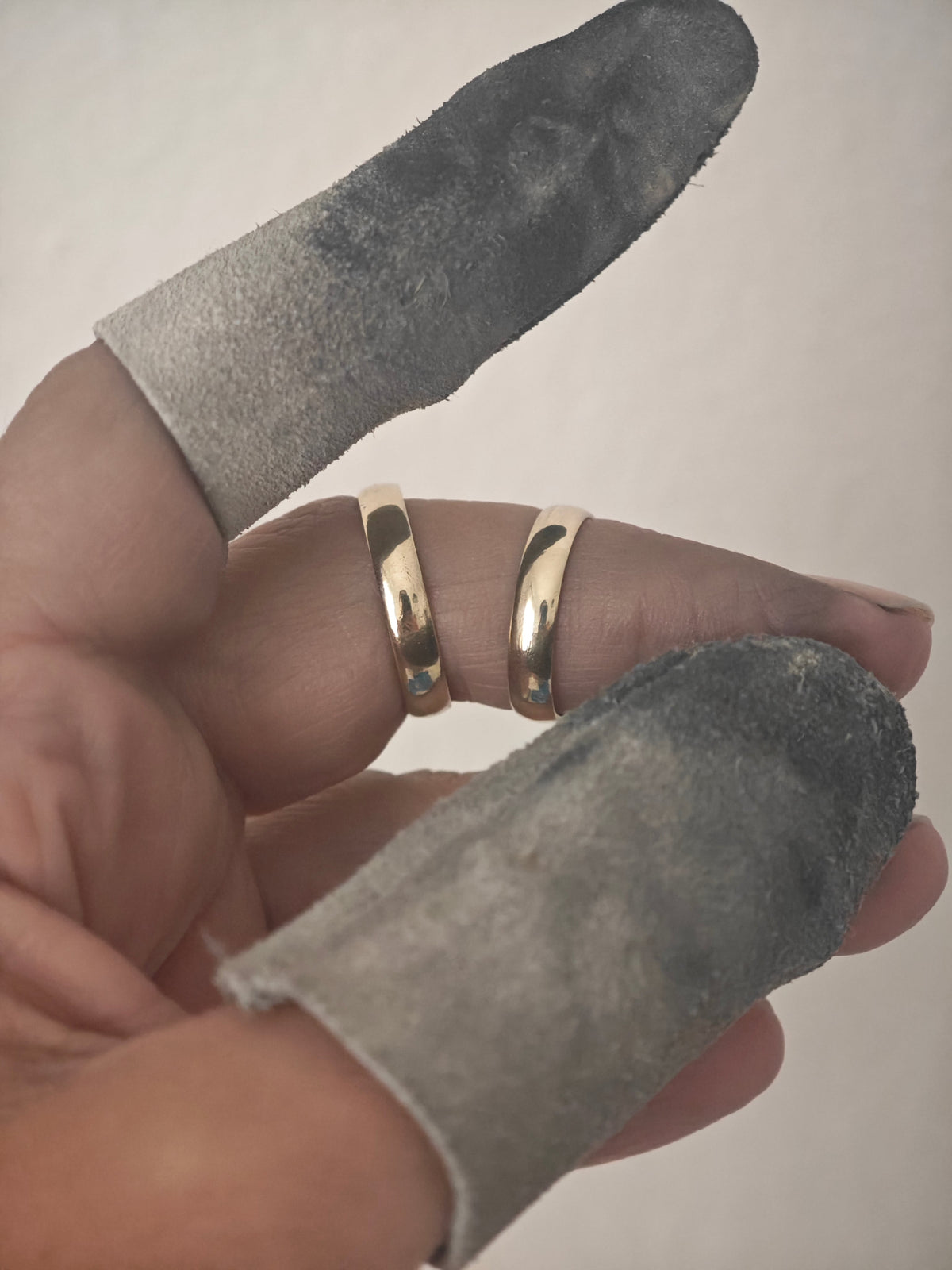 A close-up of a hand wearing two slim gold rings, with gray fabric finger sleeves covering the fingers against a plain light background.