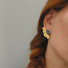 Close-up of a person wearing gold earrings with a blue gemstone against a neutral background