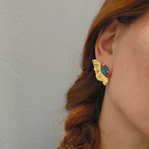 Close-up of a person wearing gold earrings with a blue gemstone against a neutral background