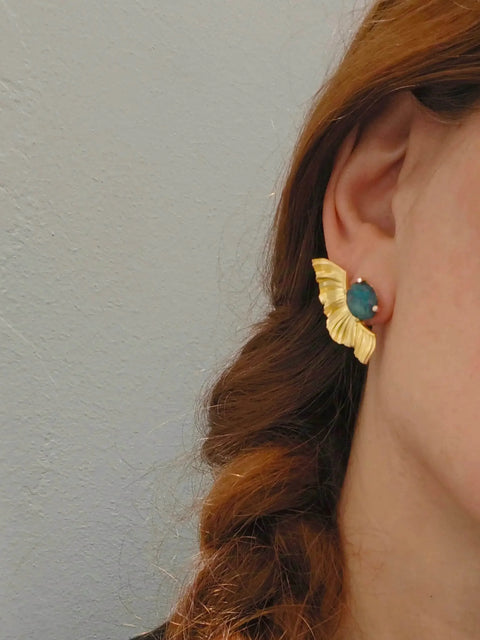 Close-up of a person wearing gold earrings with a blue gemstone against a neutral background