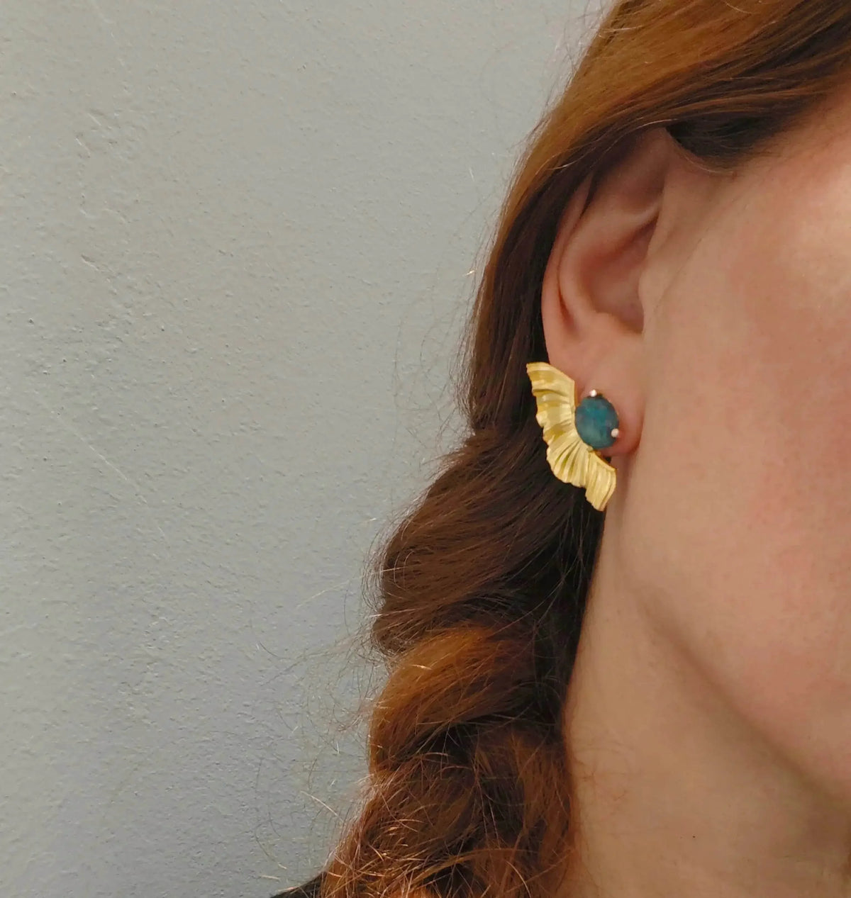 Close-up of a person wearing gold earrings with a blue gemstone against a neutral background