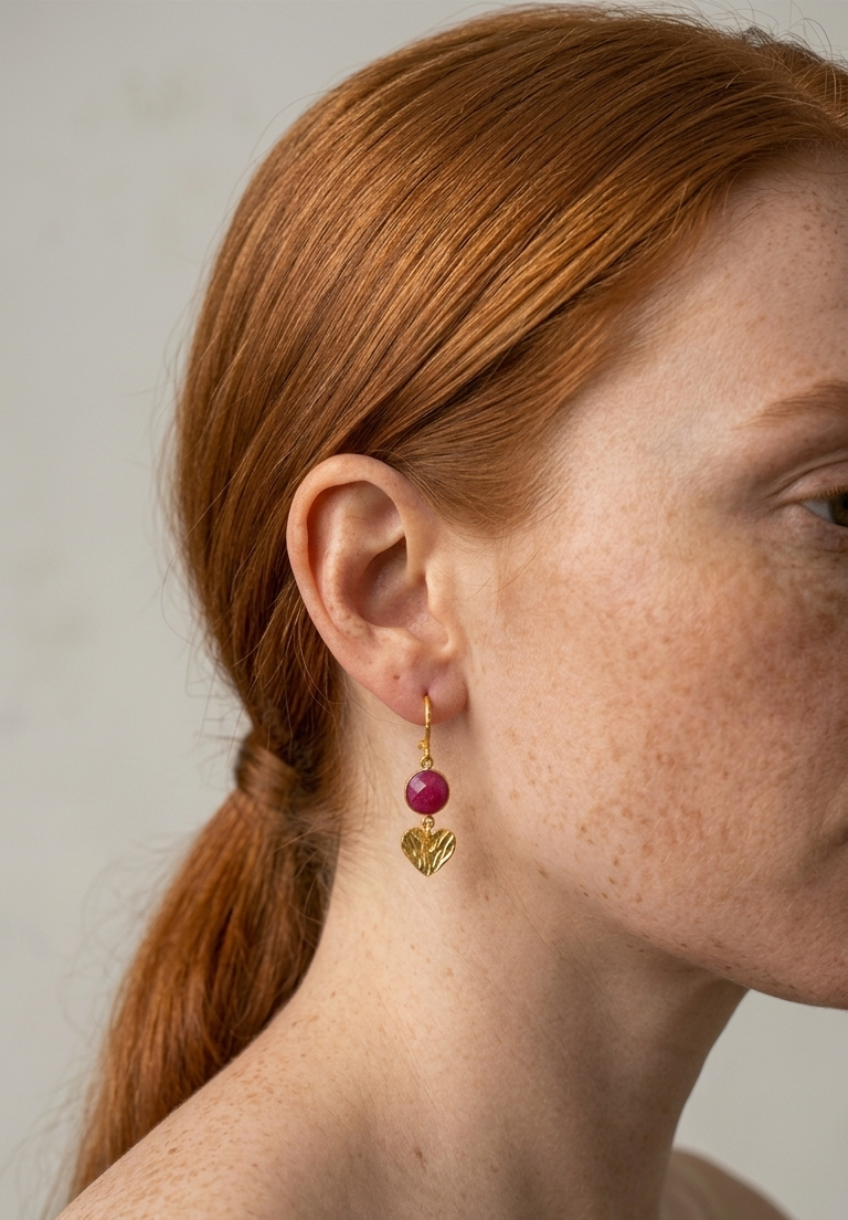 Close-up of a person wearing gold love ( heart ) earrings with a ruby  stone by Athenart made in Athens 