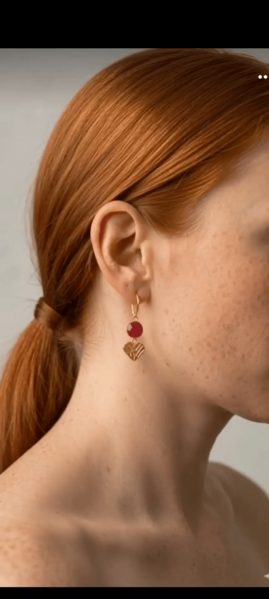 Close-up of a person wearing gold  heart (love) earrings with red ruby stones by Athenart, made in Athens 