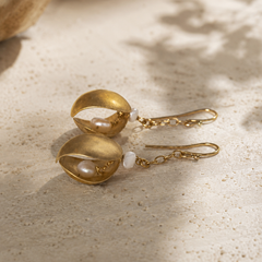 A pair of gold drop earrings with textured gold leaf-like cups holding small white pearls, resting on a textured beige stone surface with soft shadows.