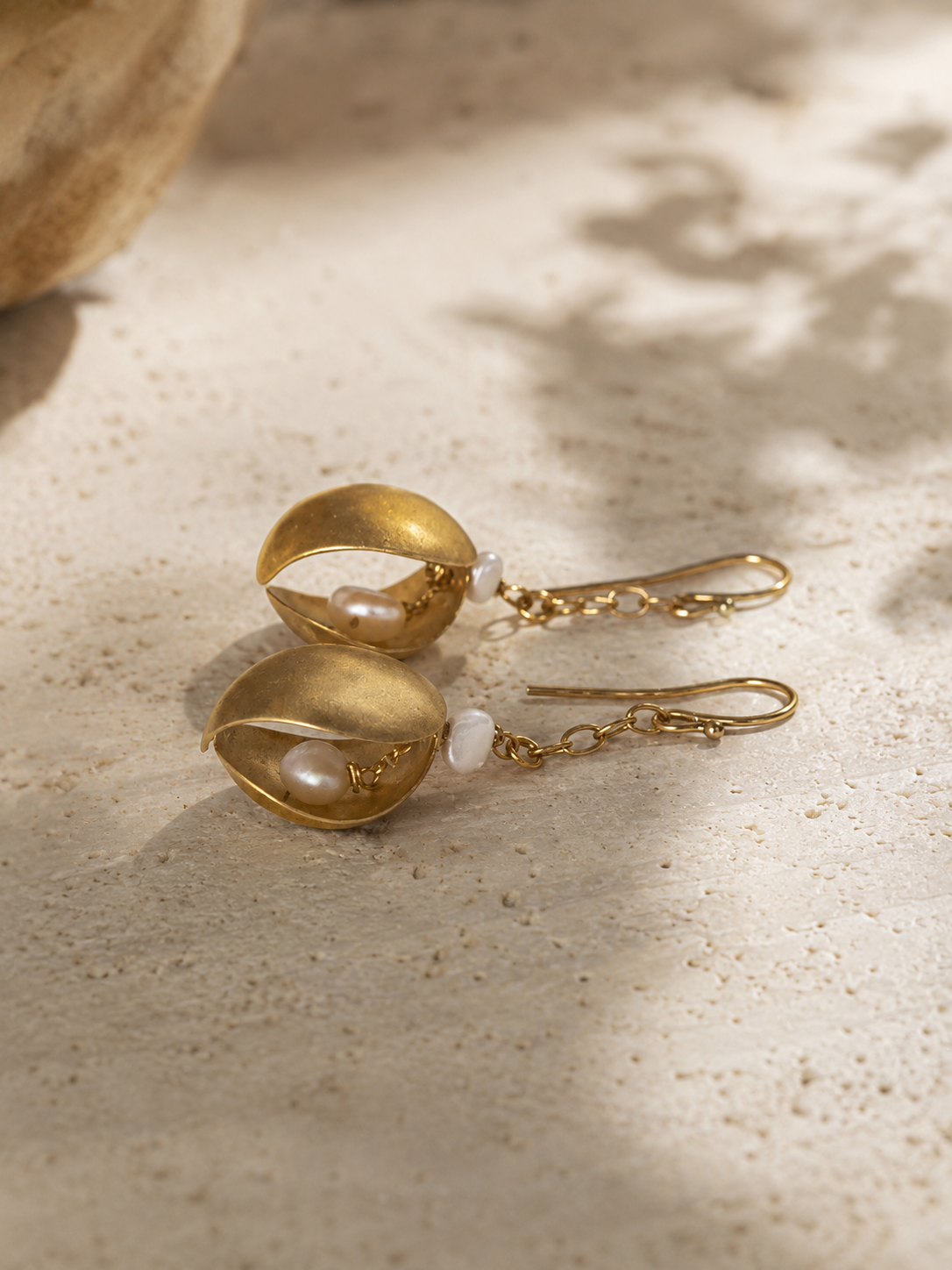 A pair of gold drop earrings with textured gold leaf-like cups holding small white pearls, resting on a textured beige stone surface with soft shadows.