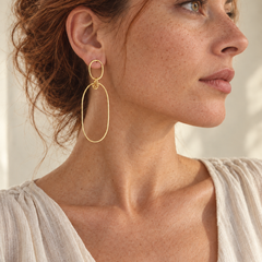 A woman with red hair wearing a delicate gold drop hoop earring, dressed in a light beige linen top against a softly lit neutral background.
