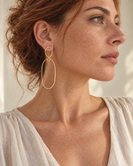 A woman with red hair wearing a delicate gold drop hoop earring, dressed in a light beige linen top against a softly lit neutral background.