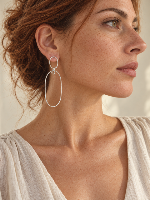 A close-up side profile of a woman with reddish-brown hair wearing a delicate silver double-ring hoop earring. She is wearing a light beige textured top against a softly lit, warm neutral background.