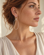 A close-up side profile of a woman with reddish-brown hair wearing a delicate silver double-ring hoop earring. She is wearing a light beige textured top against a softly lit, warm neutral background.