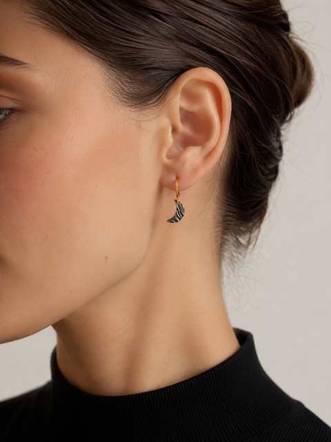 Brunette model wearing a small crescent moon earring with a gold hoop and dark textured charm, shown in a close-up side profile against a soft neutral background.