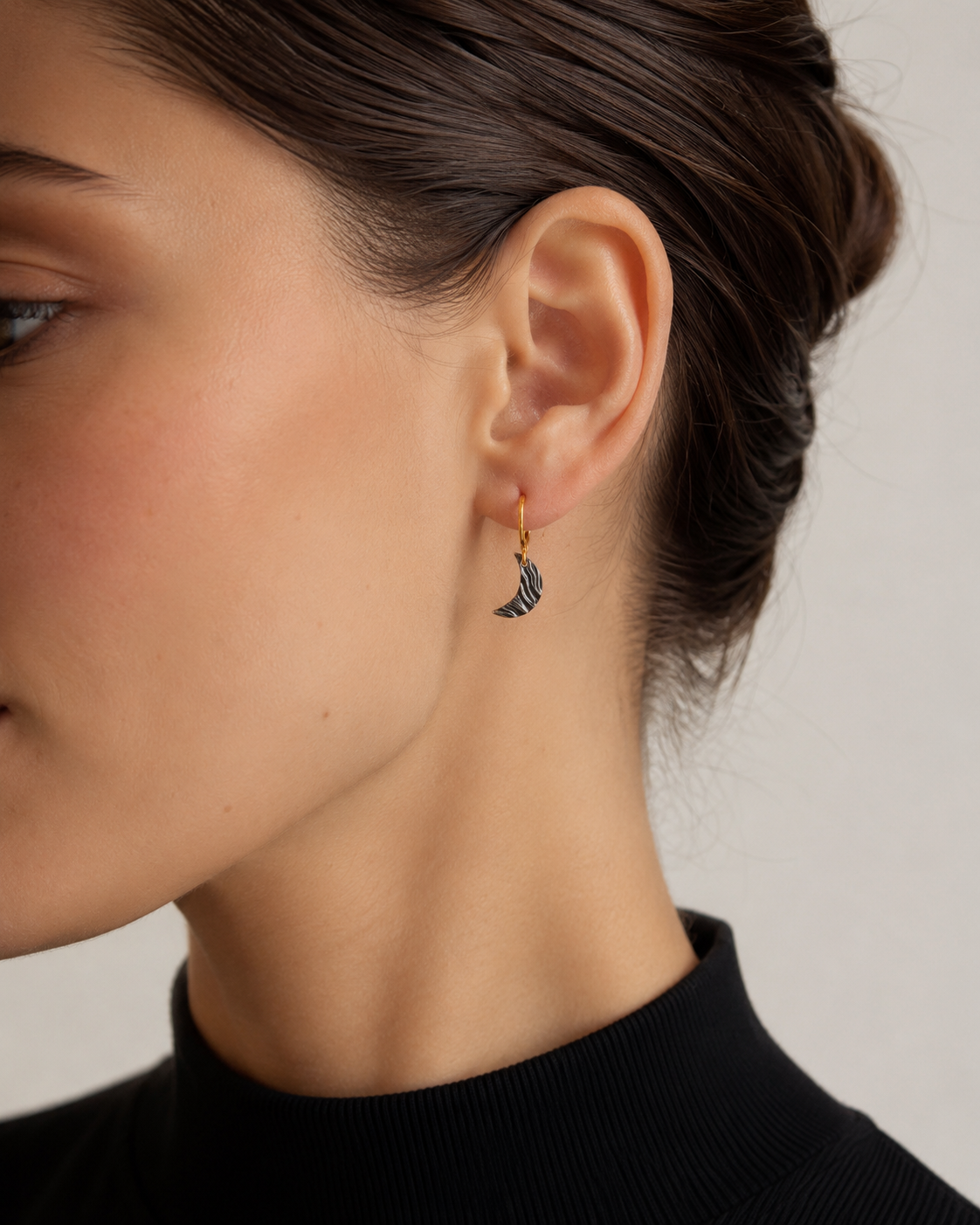 Brunette model wearing a small crescent moon earring with a gold hoop and dark textured charm, shown in a close-up side profile against a soft neutral background.