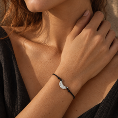 A close-up lifestyle shot of a woman wearing a delicate black cord bracelet with a silver crescent moon charm, set against a warm, sunlit background.