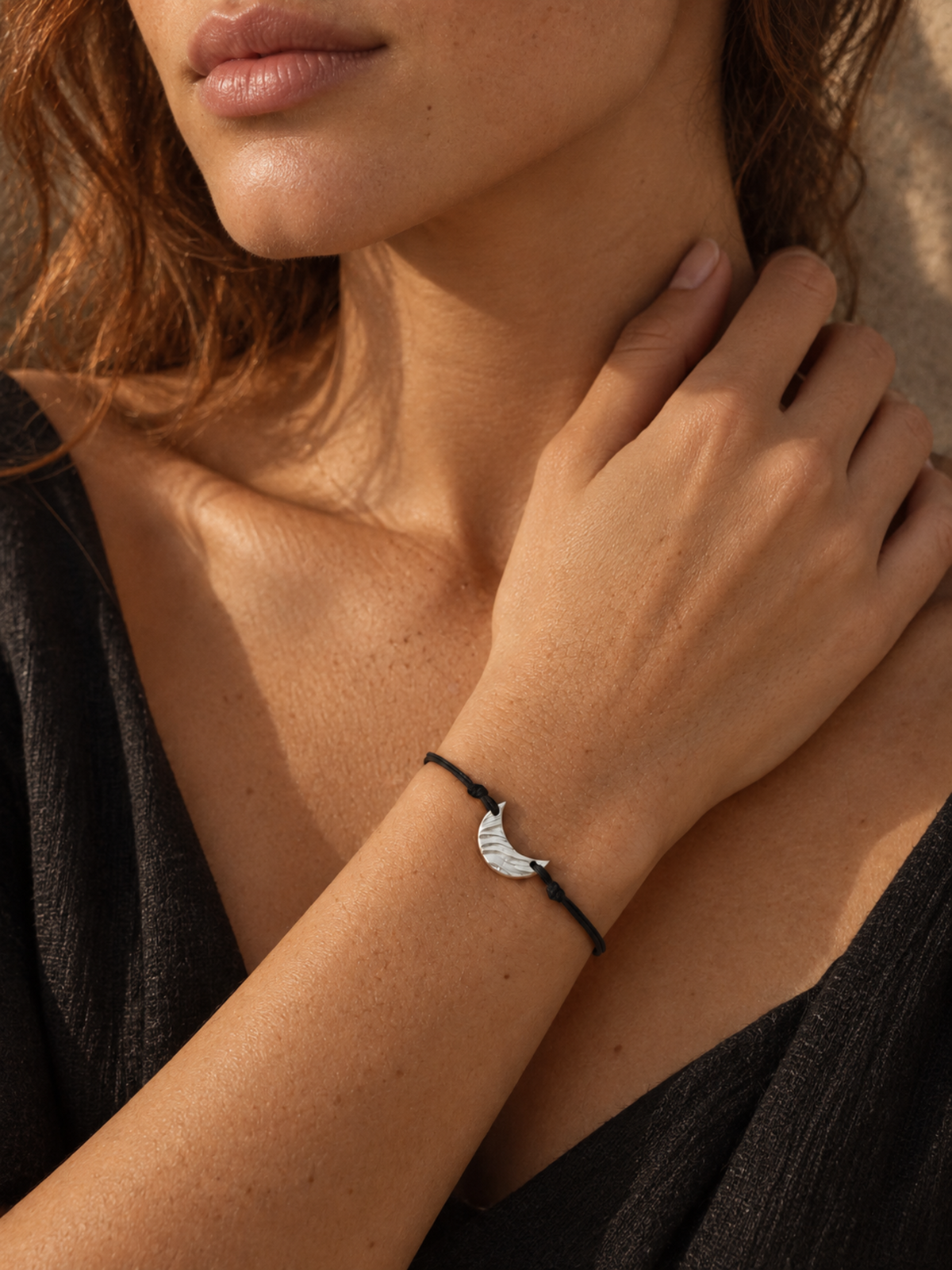 A close-up lifestyle shot of a woman wearing a delicate black cord bracelet with a silver crescent moon charm, set against a warm, sunlit background.